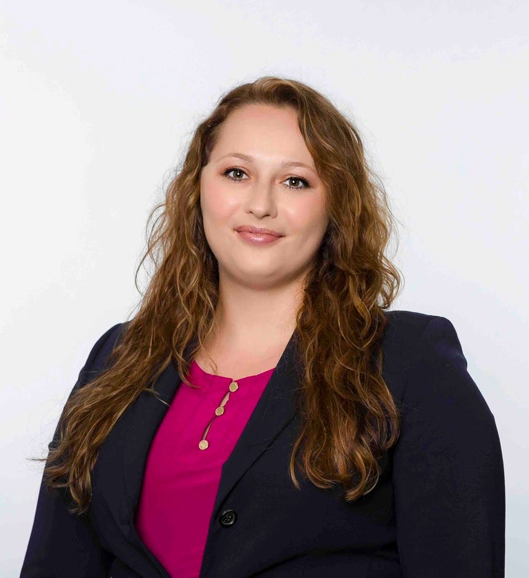 This photo pictures School of Humanities and Arts Event Specialist, Kelly Franks, in a navy blazer with a bright fuscia blouse underneath. She is standing in front of a white backdrop, smiling.