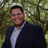 Photo of Javier Carmona wearing glasses, a blue blazer, white and blue printed button up, and a tree landscape behind him.