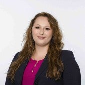 This photo pictures School of Humanities and Arts Event Specialist, Kelly Franks, in a navy blazer with a bright fuscia blouse underneath. She is standing in front of a white backdrop, smiling.